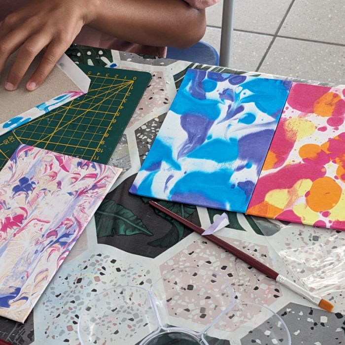 Mains d'enfant appliquant de la colle sur un rembord de sa couverture, à côté de couvertures déjà contrecollées