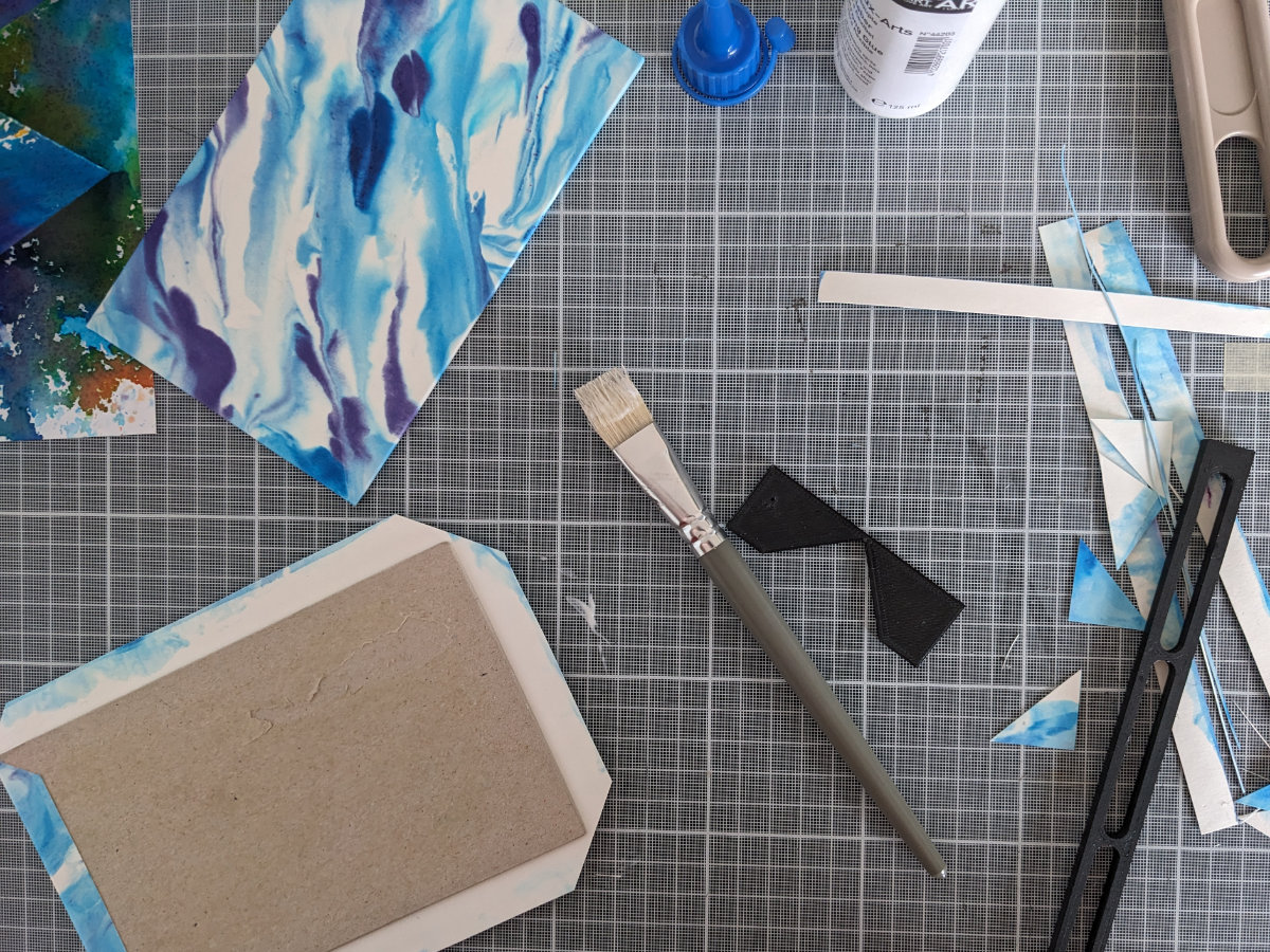 Aperçu de l'espace de travail lors du contrecollage des couvertures : papier marbré, carton gris, colle, pinceau, outils de découpe...